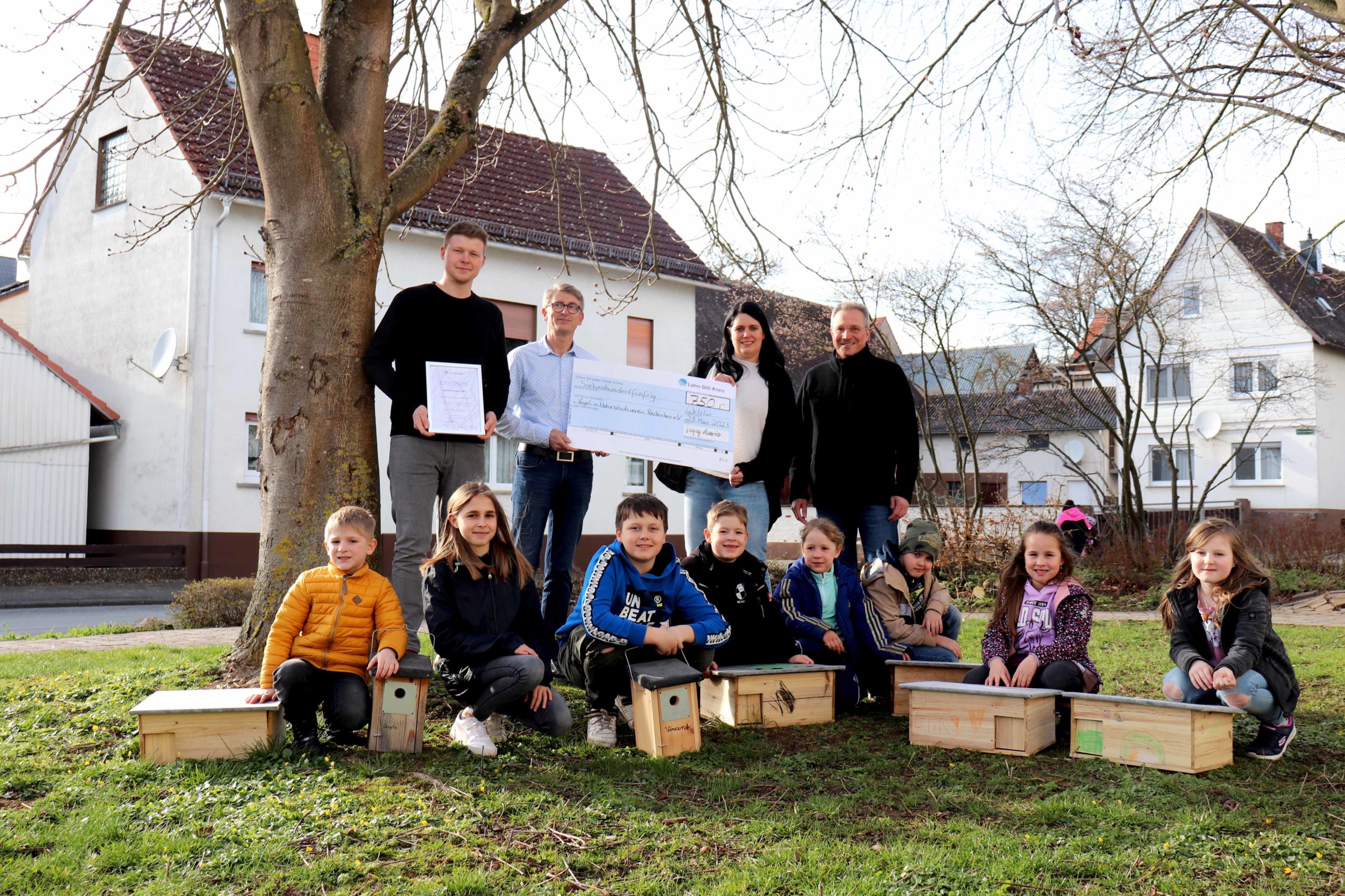 Martin Magnus und Fabian Olbrich vom Vogel- und Naturschutzverein Reiskirchen freuen sich gemeinsam mit Kreis-Umweltdezernentin Andrea Biermann und Hüttenbergs Bürgermeister Christof Heller über das Preisgeld, mit dem ein großes Insektenhotel in der Gemeinde entstehen soll. Andrea Biermann übergibt einen großen Scheck an Martin Magnus. Im Vordergrund sitzen Kinder des Vereins mit selbstgebauten Vogelnistkästen und Igelhütten. Foto: Lahn-Dill-Kreis