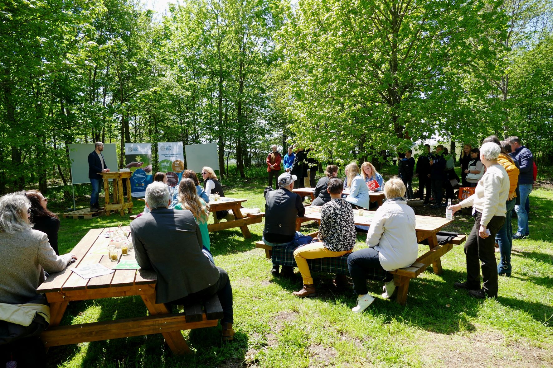 Das "Waldklassenzimmer" ist eine Fläche im Wald mit Holzstühlen und -tischen. Die verschiedenen Akteurinnen und Akteure der Preisverleihung sind im Waldklassenzimmer verteilt und unterhalten sich miteinander.