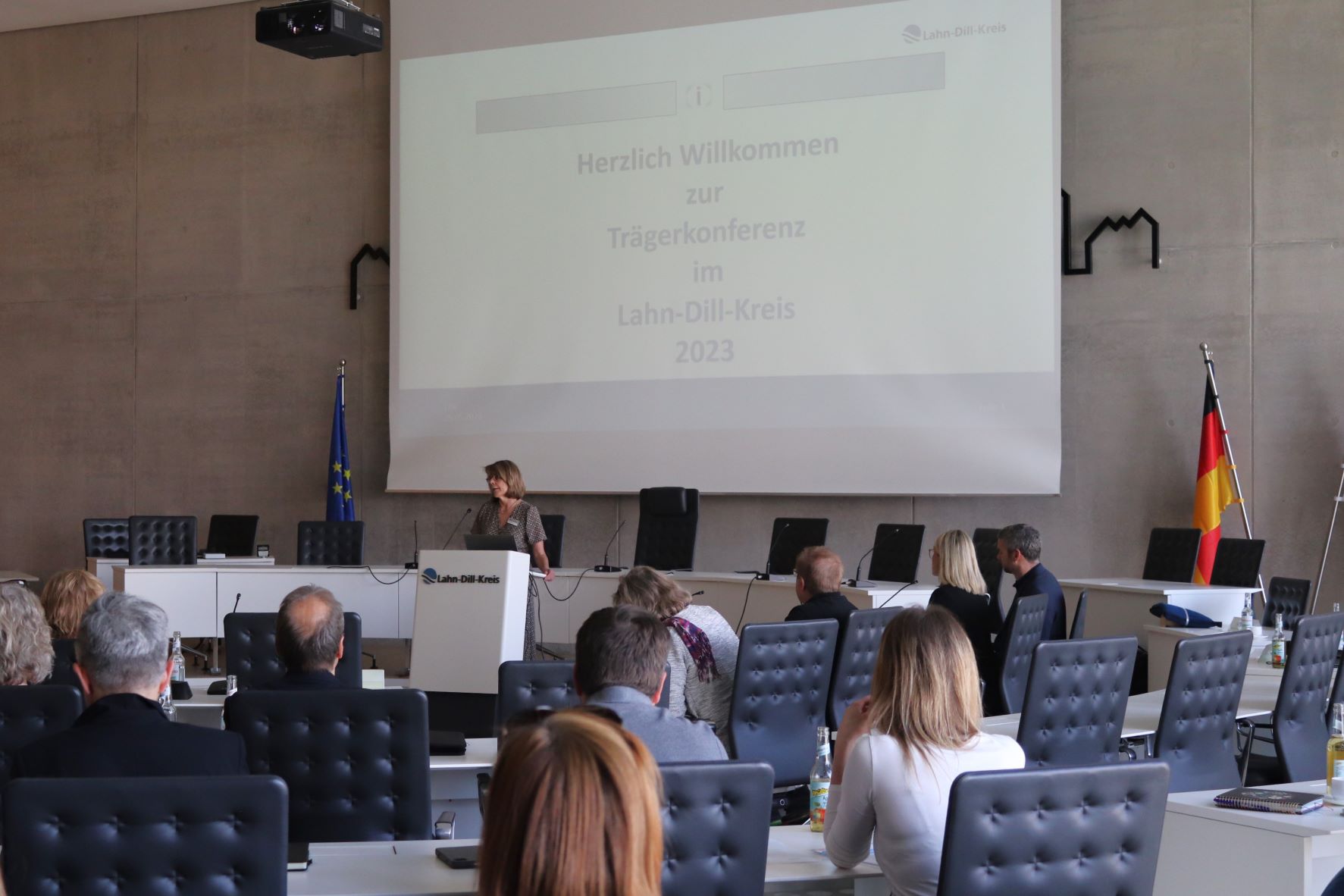 Anke Brommont-Schmidt steht am Rednerpult im Kreistagssitzungssaal. Hinter ihr ist auf einer Leinwand die Präsentation für die Konferenz sichtbar. Vor ihr sitzen einige Menschen, die ihrer Rede zuhören.