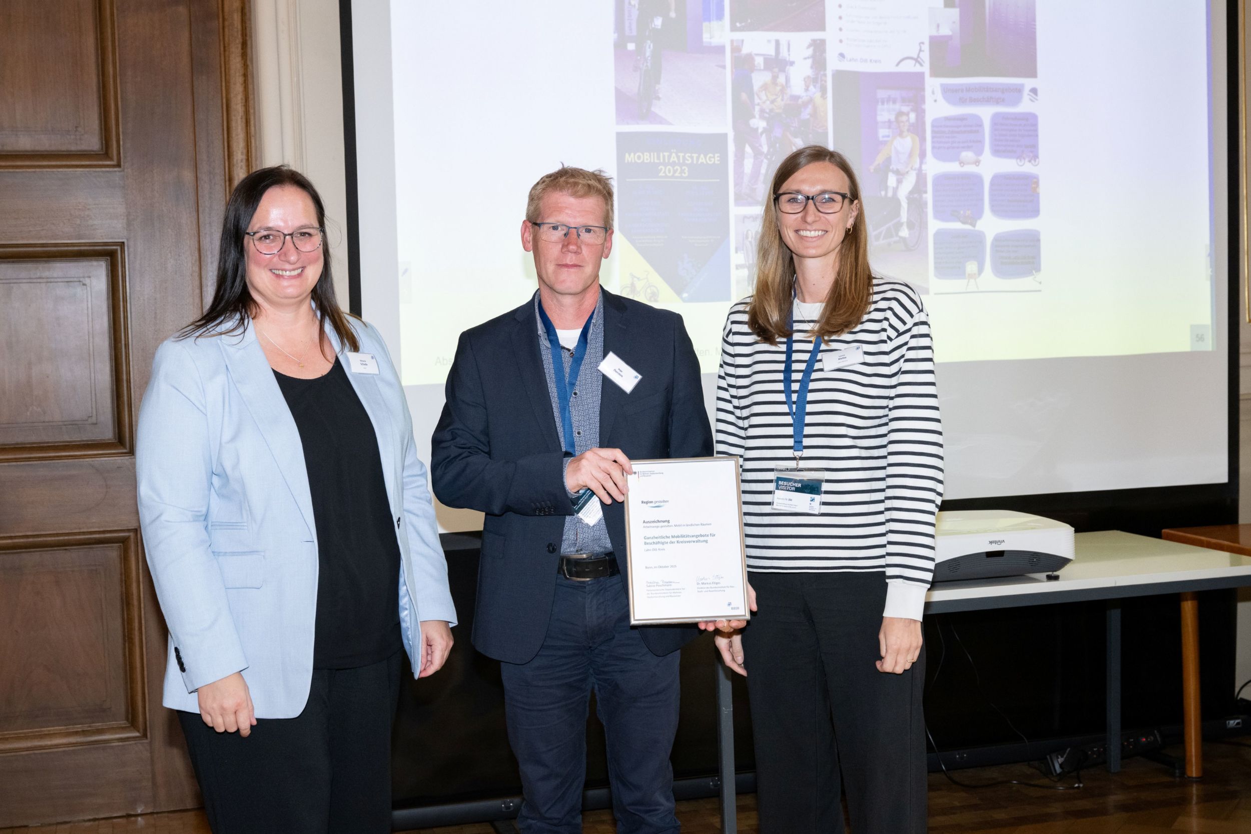 215_Auszeichnung Arbeitswege gestalten. Mobil im ländlichen Raum_Foto BBSR Ingo Dorsten (Mitte) und Laura Mette (rechts), die das Mobilitätskonzept der Kreisverwaltung umgesetzt haben, nahmen die Auszeichnung von Melanie Schade, Leiterin des PG Kompetenzzentrums für Ländliche Mobilität (KoLMo) (links) entgegen. Foto: BBR/BBSR/bundesfoto/BA
